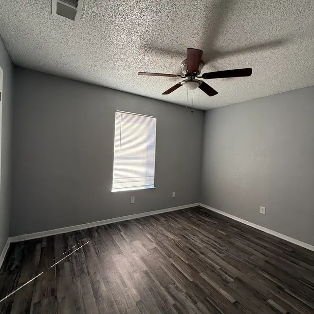 A spacious, empty room with gray walls and a ceiling fan. It has a window with white blinds allowing natural light to enter the space. The floor features dark wood-like laminate. 