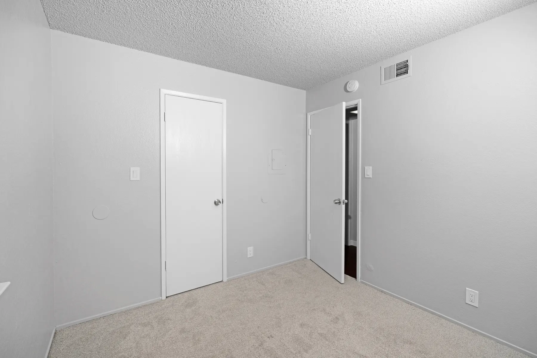 A small, empty room with light gray walls and beige carpet. There are two doors on the right side, one white and one darker, and the room is well-lit with a textured ceiling.