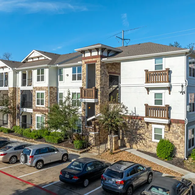 A modern apartment complex featuring three stories with a mix of stone and siding on the exterior. Several cars are parked in front on a paved lot with greenery surrounding.