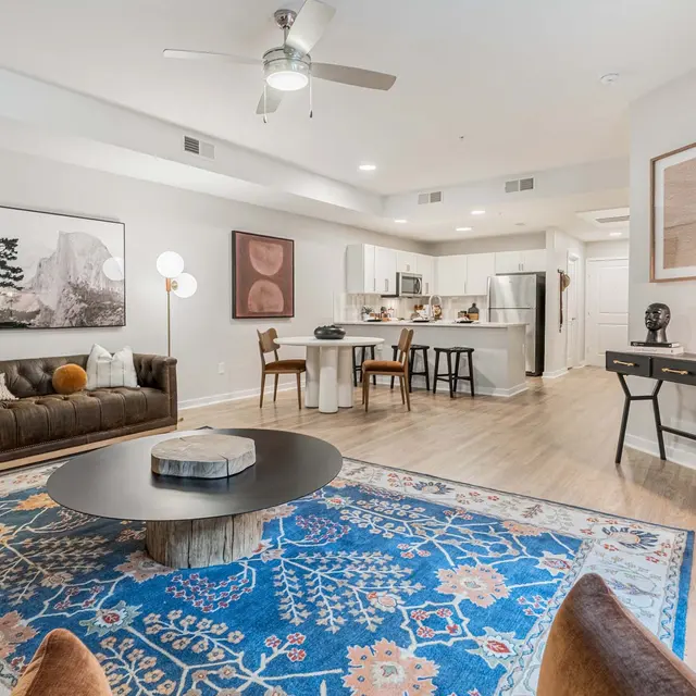 A modern living room featuring a leather sofa, a large area rug with intricate patterns, and a coffee table at the center. The dining area is visible in the background with a kitchen adjacent to it. The space is well-lit and has decorative wall art.