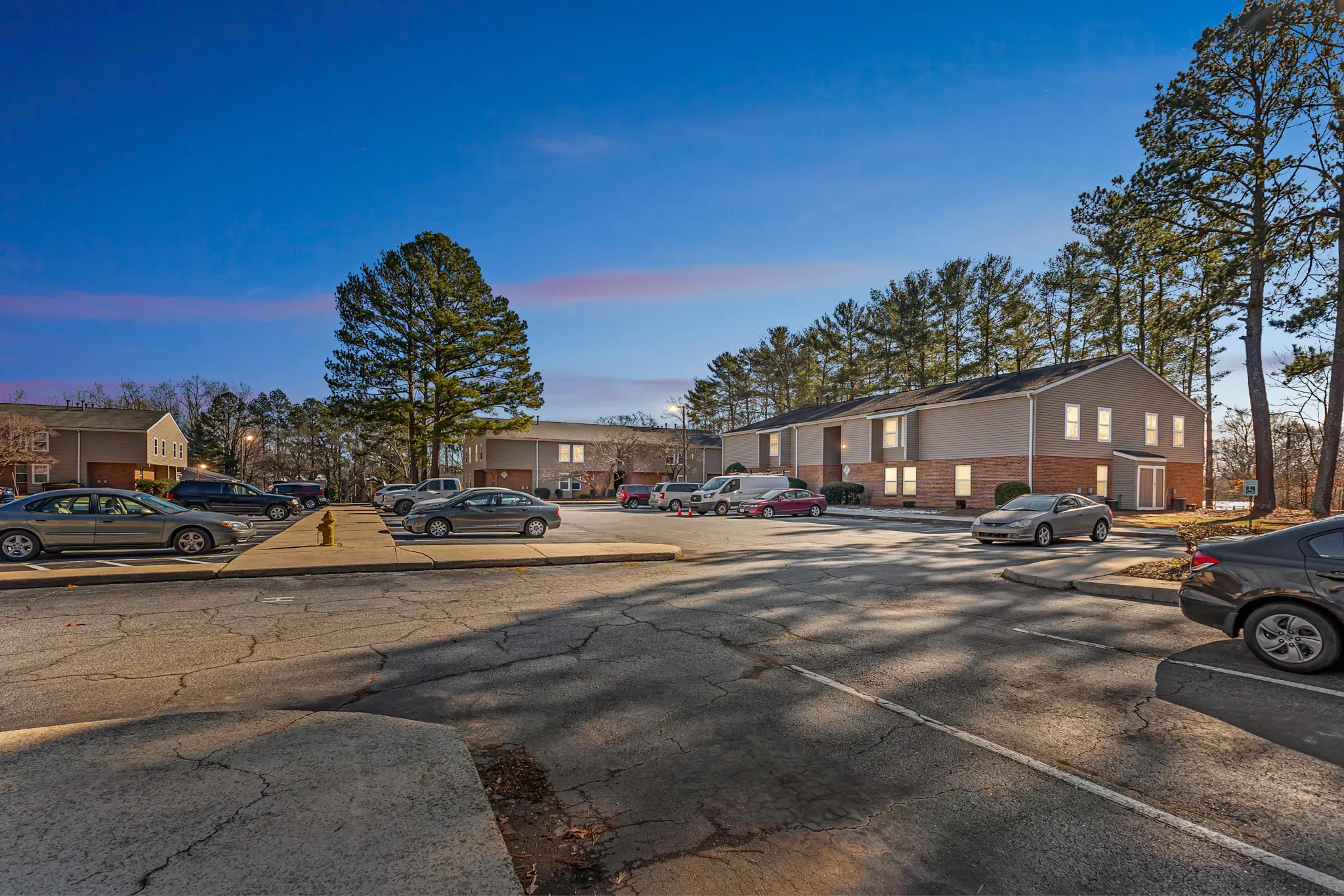 A view of an apartment complex from the parking lot, featuring several vehicles parked and trees in the background.
