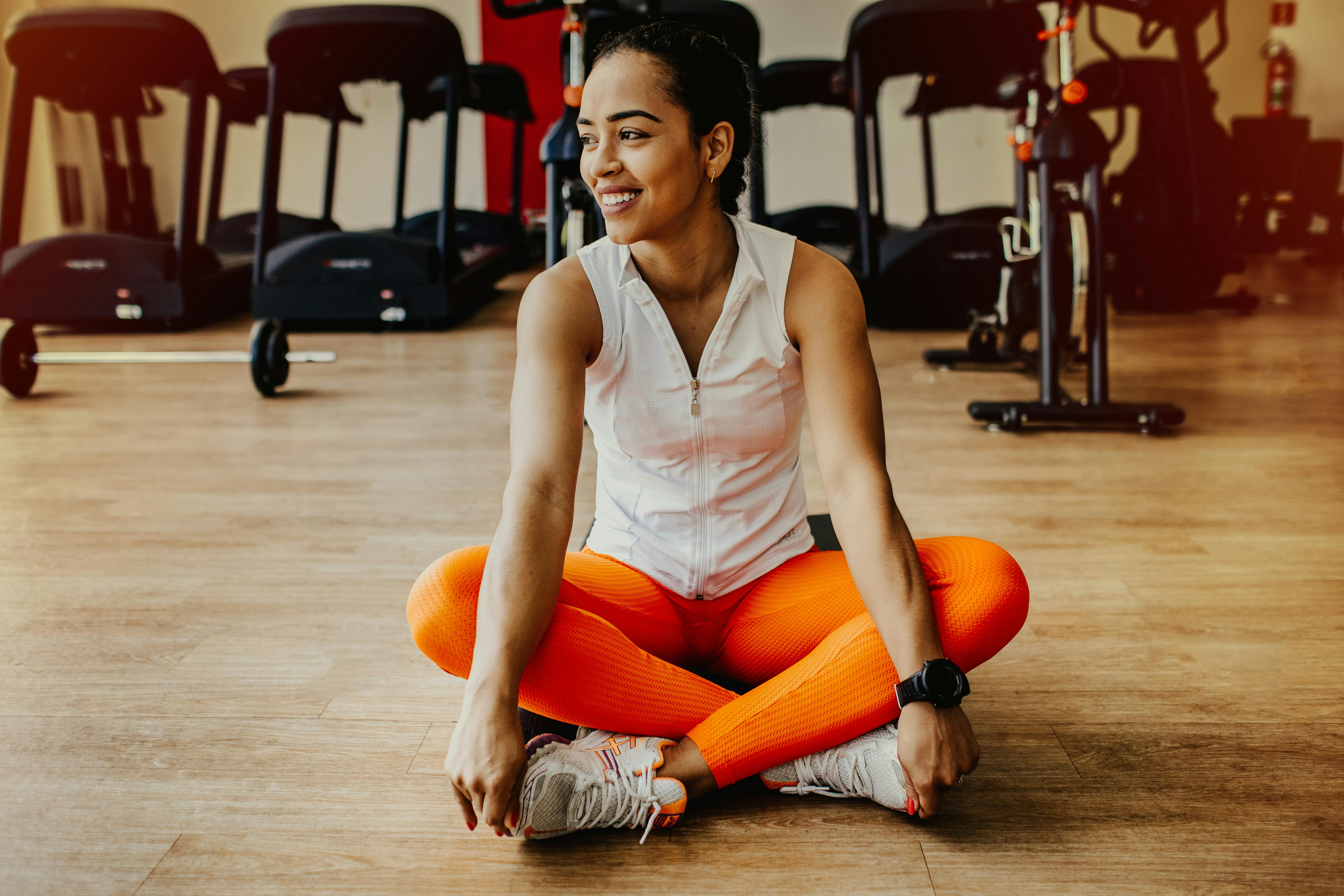 Joyful Fitness Moment A smiling woman sitting cross-legged on a wooden gym floor, wearing a white sleeveless top and bright orange leggings. She has her hair tied back and is surrounded by gym equipment like treadmills and weights in the background.