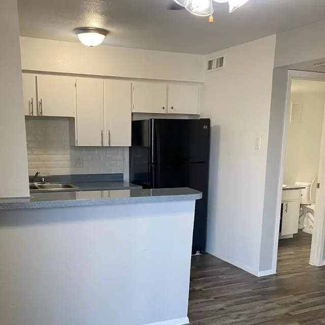 A modern kitchen featuring white cabinets, a black refrigerator, and a granite countertop. The kitchen area is partially open to the living space with a view of a bathroom door in the background.