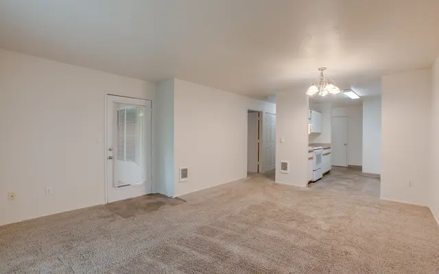 A spacious living room with light beige carpeting and white walls. On the left, there's a door leading outside. The right side features an entrance to the kitchen area, which includes a white refrigerator and closed cupboards, and a door leading to another room.