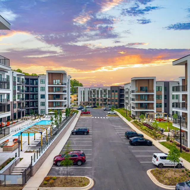 A modern apartment complex with multiple buildings, a swimming pool area, and a landscaped setting at sunset.