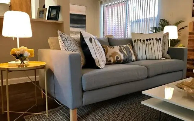 A cozy modern living room featuring a grey sofa with patterned throw pillows, a round side table with a lamp and flowers, and a large window with sheer blinds.