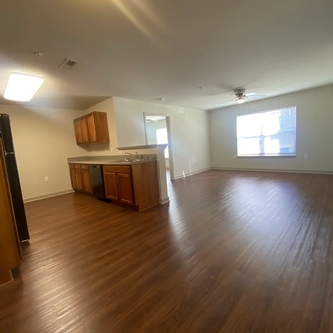 An empty living area with wooden flooring, a kitchen area featuring wooden cabinets and an open layout leading to a windowed room.
