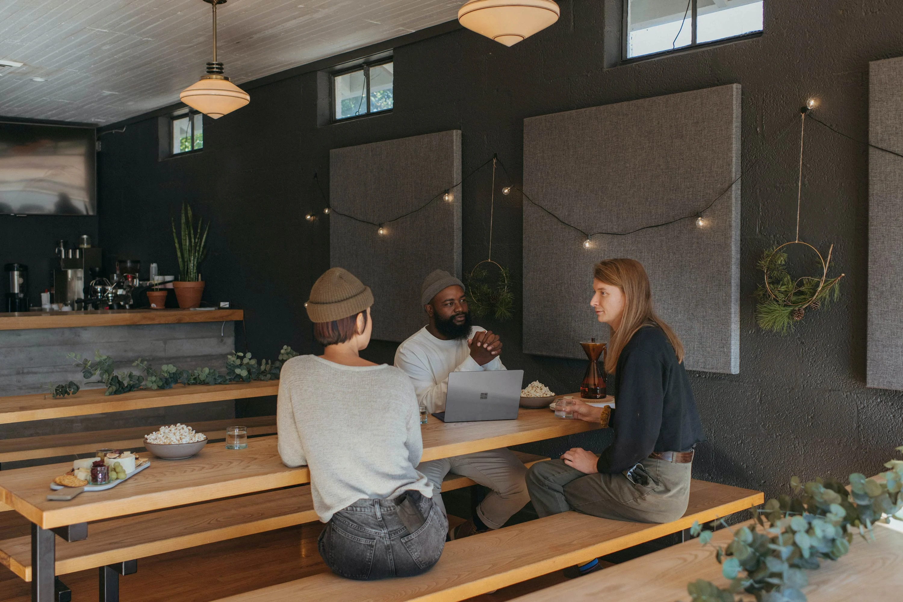 A relaxed meeting in a cafe setting with three individuals discussing at a wooden table. The interior is decorated with greenery and has a warm ambiance.
