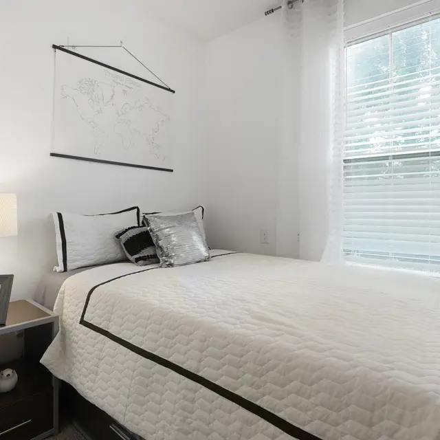 A cozy bedroom with a white bedspread, large window with sheer curtains, a bedside table with a lamp, and a wall-mounted map.
