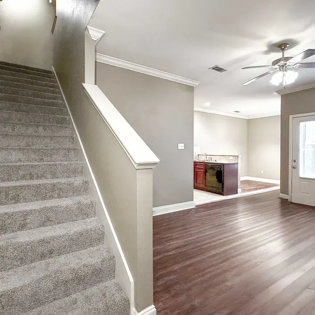 Interior view of a modern home, featuring a carpeted staircase, an open living space with hardwood flooring, and a kitchen area in the background.