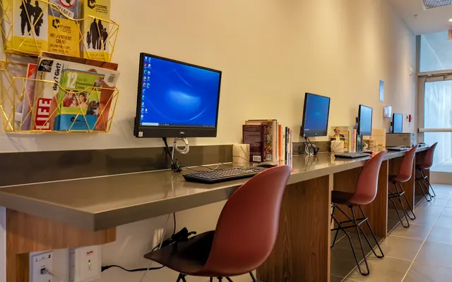 A modern workspace featuring a long counter with several computer stations, each with a monitor and keyboard. A wall-mounted rack is filled with magazines, and books are placed on the counter. There are stylish chairs at the stations, and a glass door leads to a potential entry.