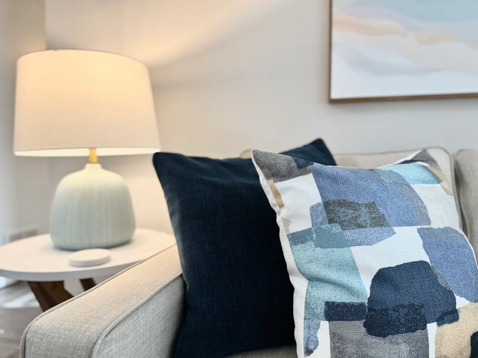 A cozy living room scene featuring a light-colored sofa with decorative pillows, a soft blue lamp, and a piece of abstract art on the wall.