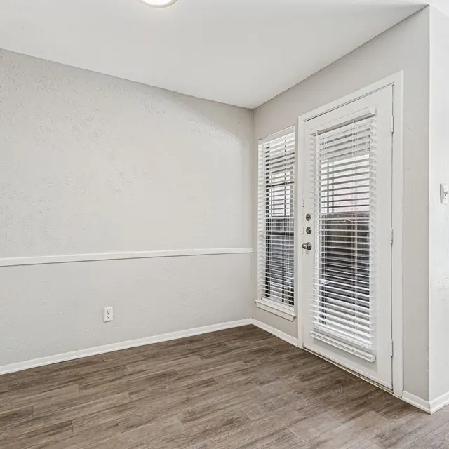 An empty corner room with light-colored walls and wooden flooring, featuring a set of patio doors with blinds.