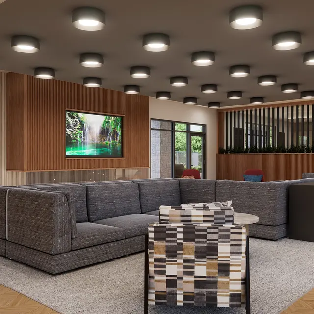 A modern lounge area featuring a large grey sectional sofa, a striped armchair, and a round coffee table. The decor includes greenery visible through large windows, a flat-screen TV on a wooden wall, and stylish ceiling lights. The flooring is light-colored wood.