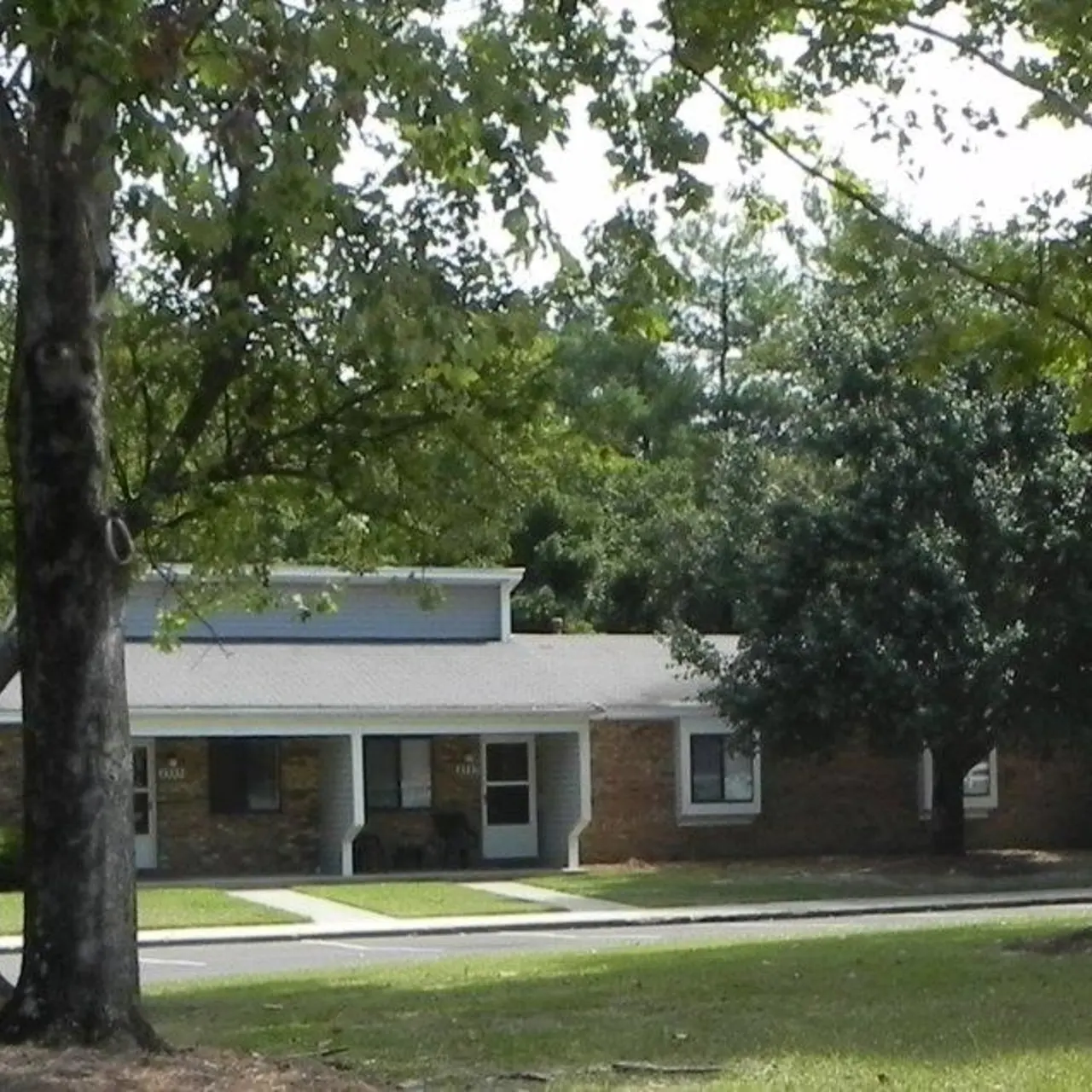 A single-story building nestled among trees, showcasing a well-kept lawn and sidewalk leading to the entrance. The building has a mix of brick and siding architecture.
