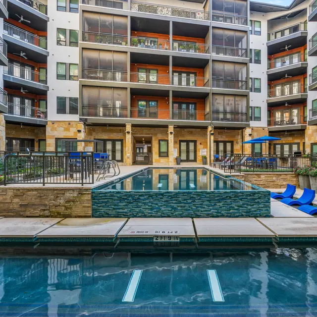A modern apartment complex with a swimming pool area. The pool is surrounded by lounge chairs and has a stone facade. The buildings are multi-storied with balconies and large windows.