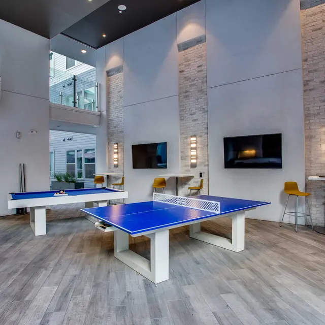 A modern game room featuring a blue ping pong table, two televisions mounted on the wall, and seating areas with yellow chairs.
