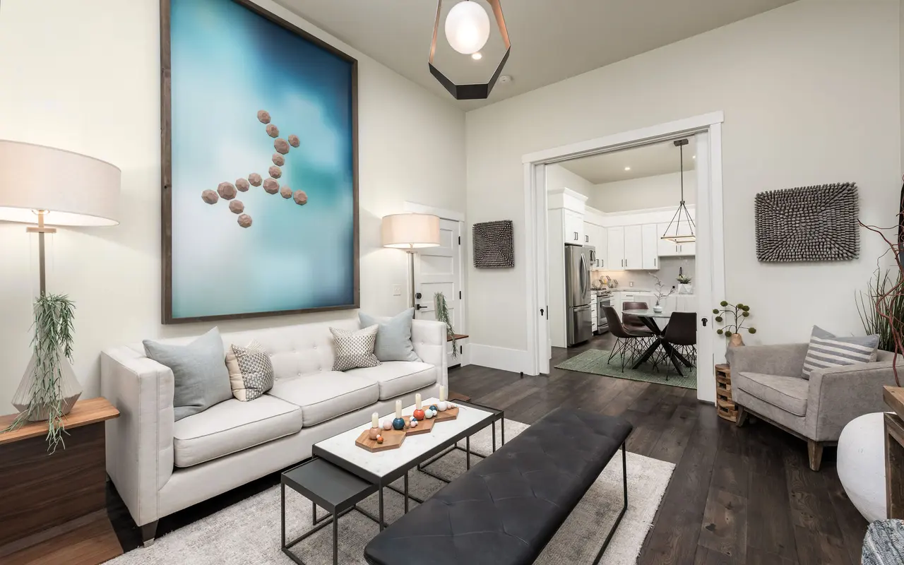 A modern living room featuring a white sofa with decorative pillows, a coffee table with natural wood decor, and a cozy armchair. The room has a large piece of abstract wall art and a bright, airy feel with natural light.