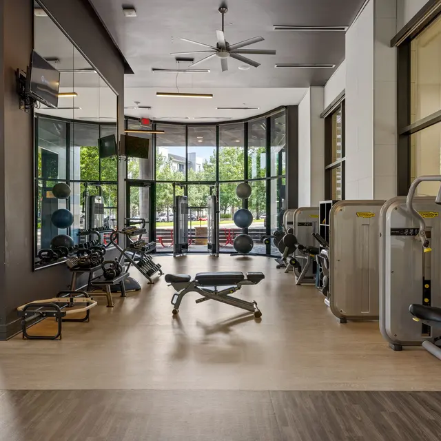 Interior view of a modern gym with large windows, fitness equipment, and hardwood flooring.