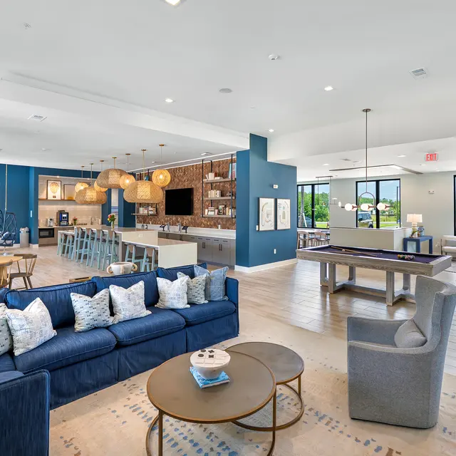 Modern Lounge and Kitchen Space A spacious modern lounge area featuring a blue sofa with decorative pillows, a round coffee table, and a dining area with wooden chairs. The kitchen is visible in the background with bar seating and a rustic vibe, illuminated by pendant lights. Large windows provide natural light and a view of the outdoors.