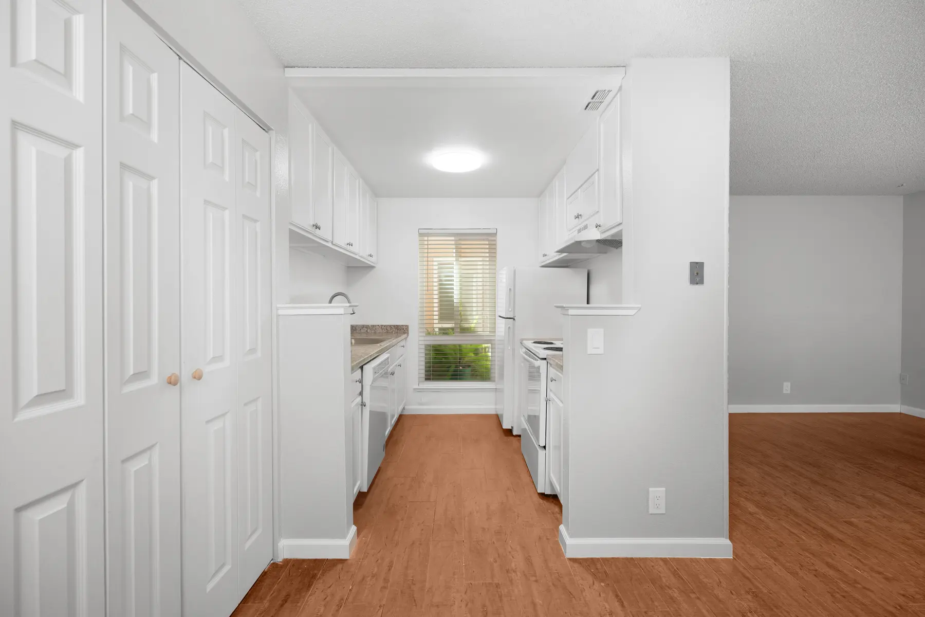 A bright, modern kitchen with white cabinetry, a window with blinds, and hardwood flooring. The kitchen features an open layout leading into a living area.