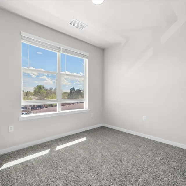 An empty room with gray carpet, light gray walls, and a window showing blue skies and clouds.