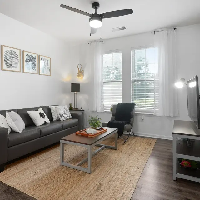 A stylish living room featuring a gray sofa, decorative pillows, a wooden coffee table with books and a plant, a dark chair, a TV unit, and large windows with natural light.