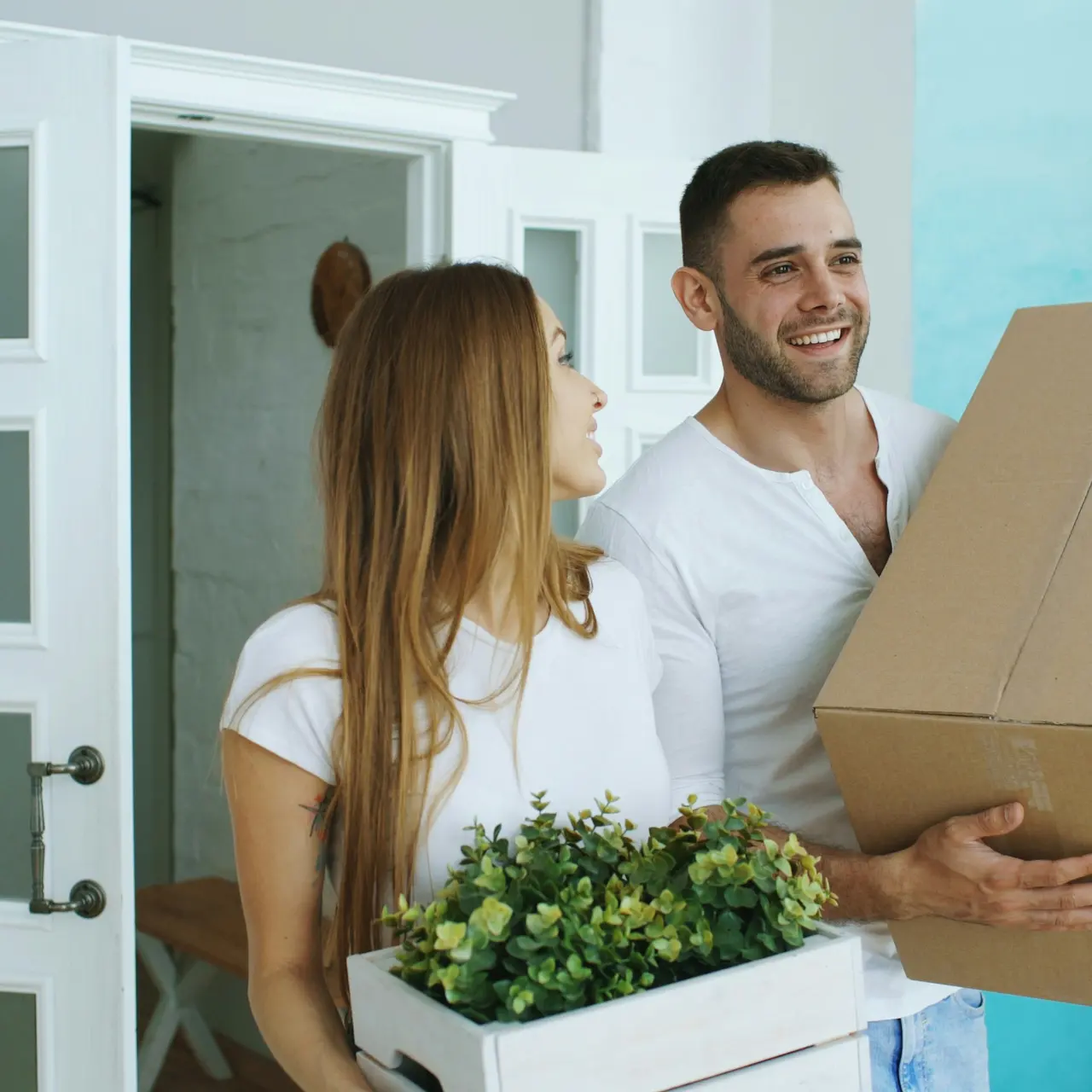 Couple Moving In A couple standing in a bright room, smiling while carrying boxes and a plant. The woman holds a box with greenery, and the man holds a large cardboard box. They are dressed casually and look happy as they move into a new space.