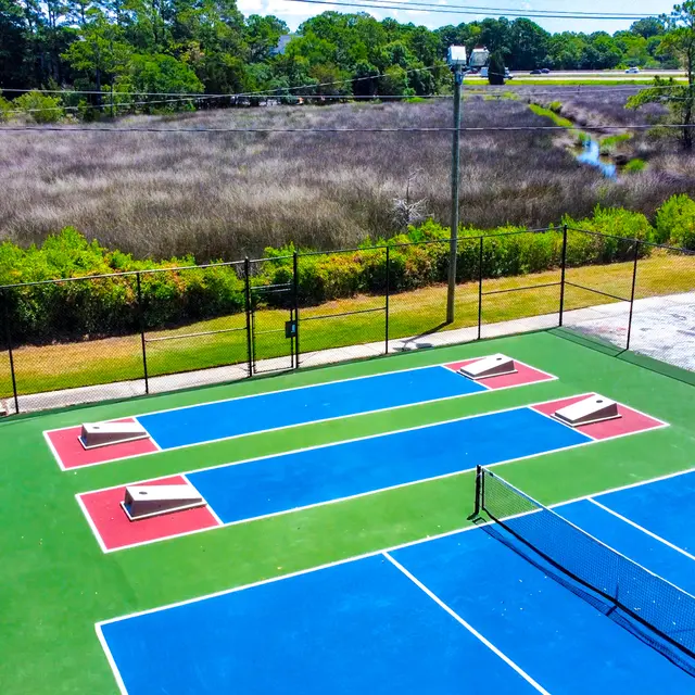 Aerial view of tennis courts with blue and red surfaces surrounded by lush greenery and a waterway in the background.