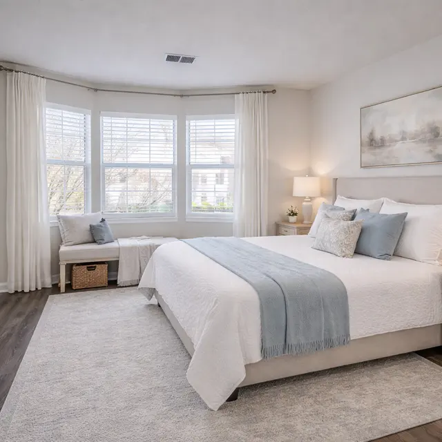 A well-decorated bedroom featuring a large bed with light bedding, large windows with sheer curtains offering natural light, and stylish bedside tables with lamps. A cozy seating area is present by the window.