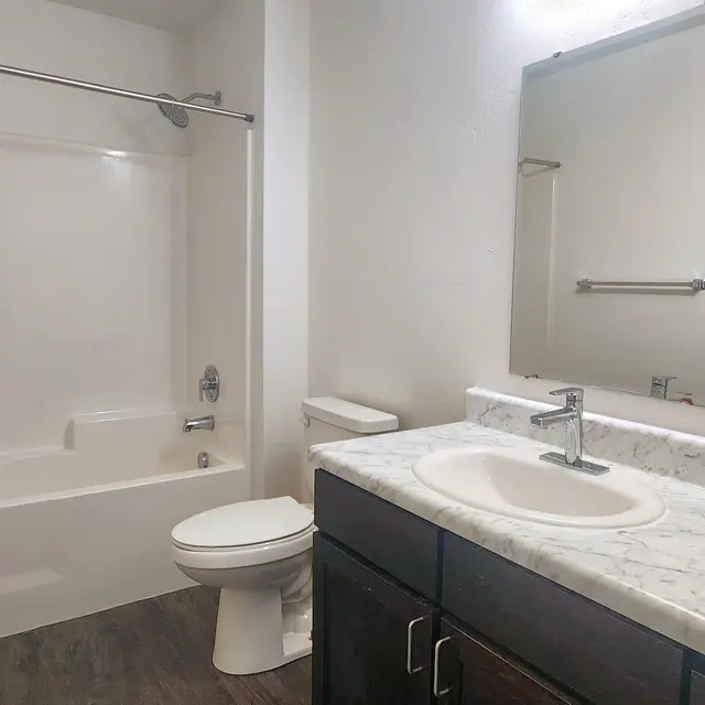 Canyon Terrace A modern bathroom featuring a white bathtub with a shower curtain, a dark wood vanity with a white marble countertop, a round sink, a large mirror, and tiled flooring.