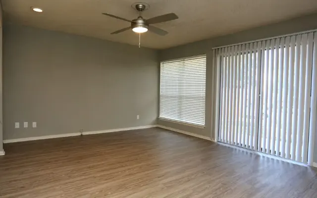 A spacious living room featuring a ceiling fan, light gray walls, and wooden flooring. There's a large window with vertical blinds allowing natural light in, and a sliding glass door that leads outside.