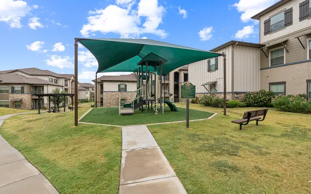 Children's Playground Area A playground area with a green shade over a play structure, surrounded by well-maintained grass and benches, set against a backdrop of apartment buildings and blue sky.