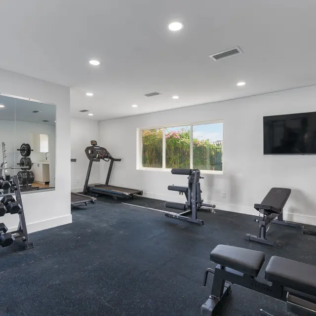 A modern home gym setup with a mirror, dumbbells on racks, a treadmill, bench press, and weight benches under bright lighting.