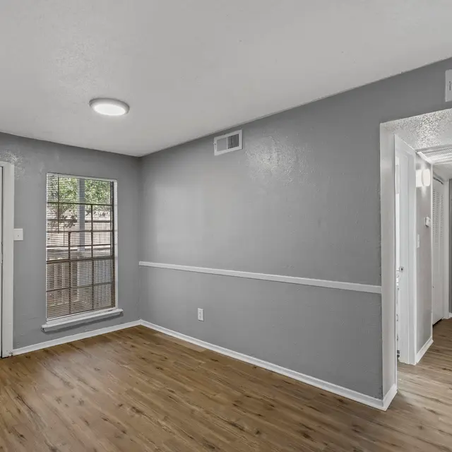 An interior view of a living room with light gray walls and wooden flooring. A front door is visible on the left, and a window is on the left wall, allowing natural light to enter. The room appears spacious with an open layout leading to a hallway visible on the right.