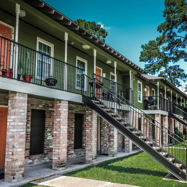 Exterior view of a two-story apartment building with orange doors and brick columns, surrounded by trees and grass.