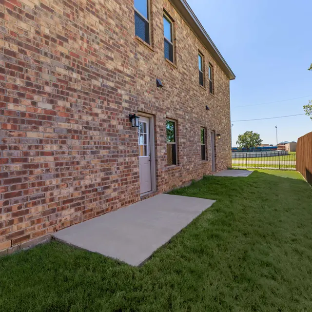 A side view of a brick building with a light-colored door, surrounded by green grass and a wooden fence.
