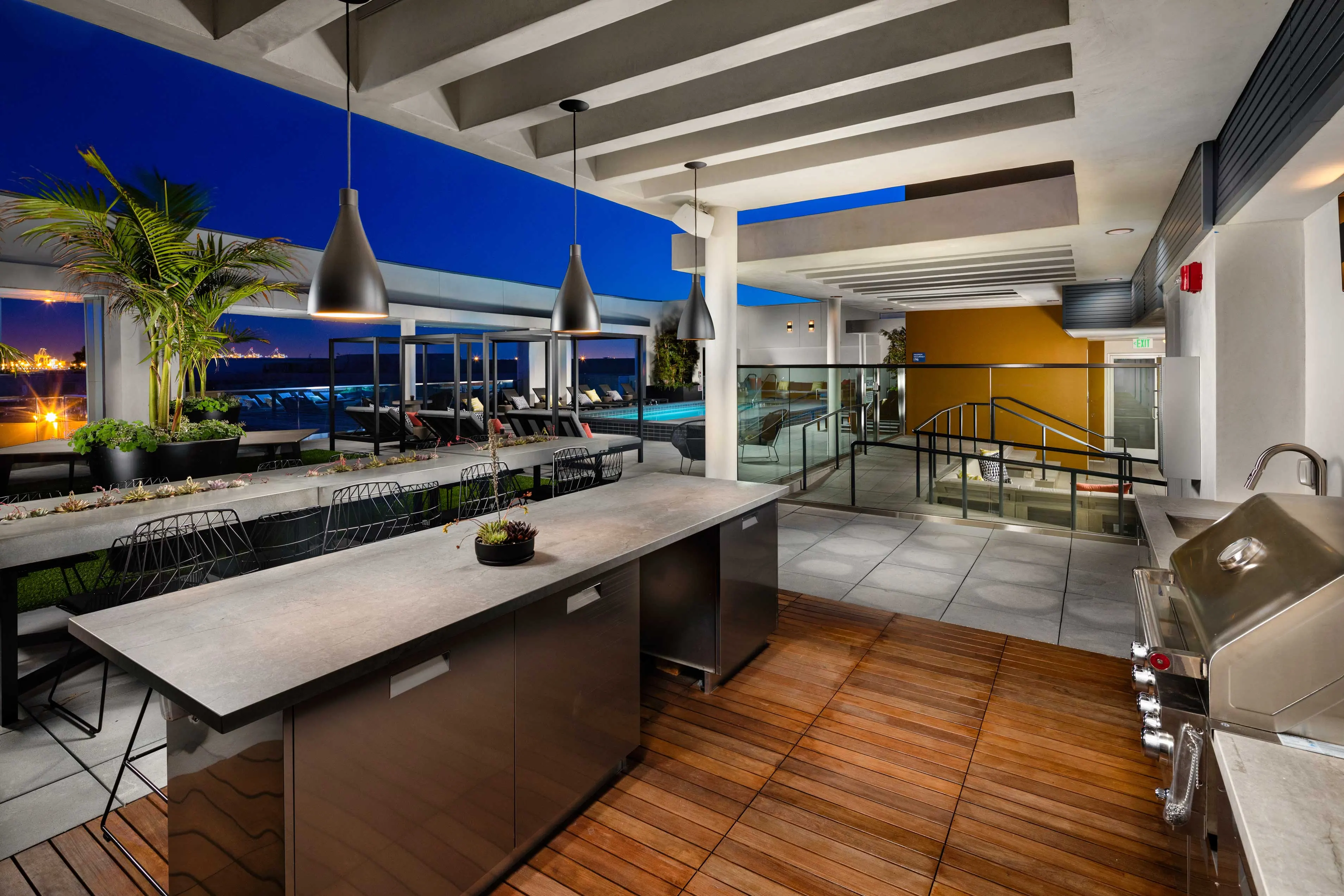 Stylish outdoor kitchen area with a large island, modern appliances, and seating overlooking a pool at dusk.