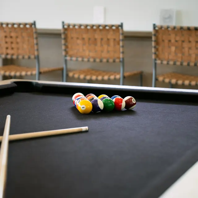 Billiard Table Ready for Play A close-up view of a billiard table featuring a set of pool balls arranged in a triangle, with two cue sticks placed on the side. In the background, there are several wooden chairs arranged in a casual setting.