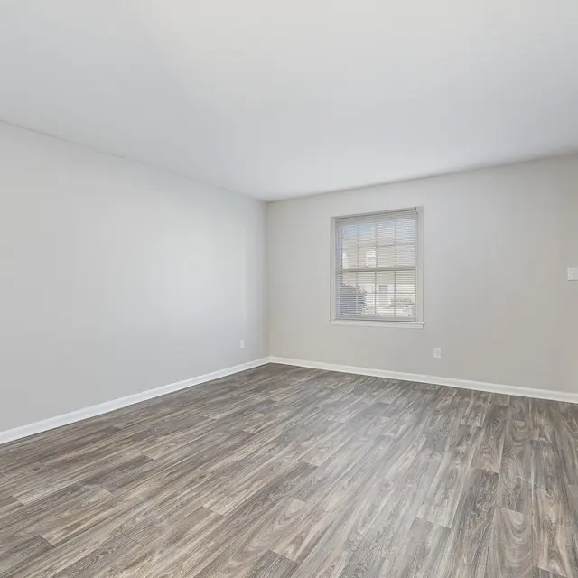 A spacious empty living room featuring light grey walls, a large window, and wood-like flooring. A staircase is visible in the corner and there’s a door on the wall to the right.