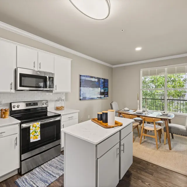 Modern Kitchen and Dining Area A modern kitchen featuring white cabinets, stainless steel appliances, and a dining area with a table and chairs. Natural light enters through the window, and a small island is visible in the center.