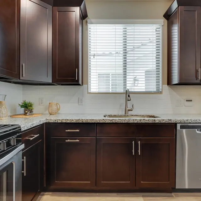 A modern kitchen featuring dark wood cabinets, stainless steel appliances, and a granite countertop. A window with blinds is visible above the sink, and there are decorative items on the counter.