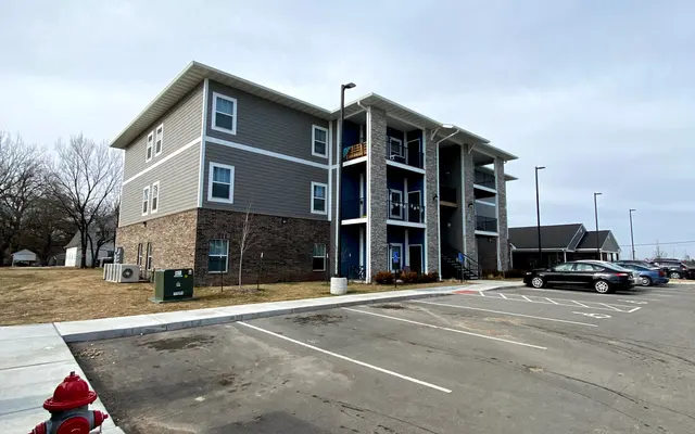 An exterior view of a three-story apartment building with gray siding and a brick bottom. The building features balconies and multiple windows. In front of the building, there is a parking lot with several empty parking spaces and a fire hydrant to the left.
