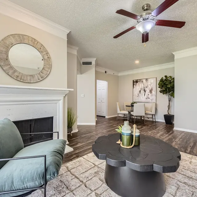 A modern living room featuring a cozy seating area with a blue-green armchair and a black coffee table, decorated with vases. A fireplace is visible, along with a leafy indoor plant and a dining area in the background.