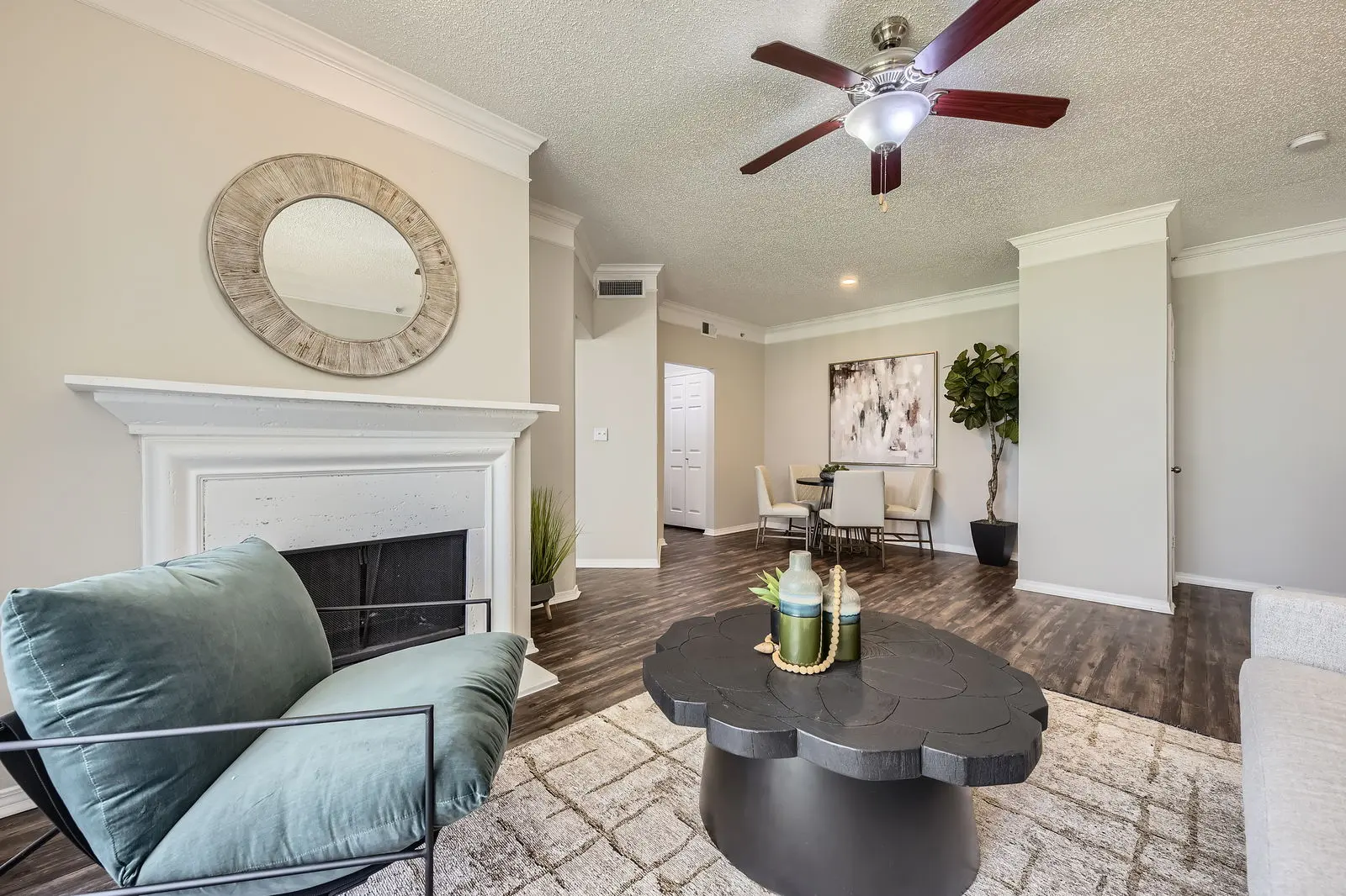 A modern living room featuring a cozy seating area with a blue-green armchair and a black coffee table, decorated with vases. A fireplace is visible, along with a leafy indoor plant and a dining area in the background.