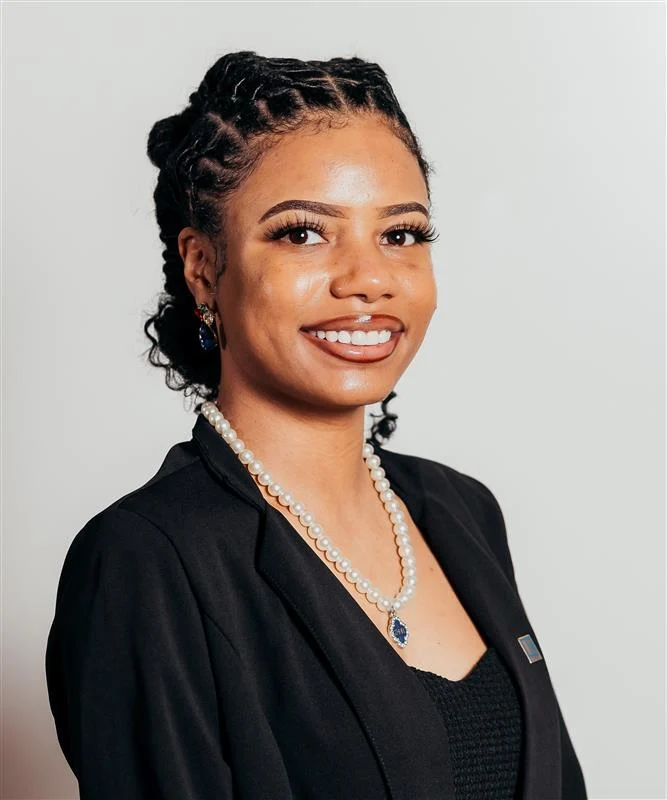 Professional Portrait of a Young Woman A smiling young woman wearing a black blazer and a pearl necklace, with braided hair styled in curls.