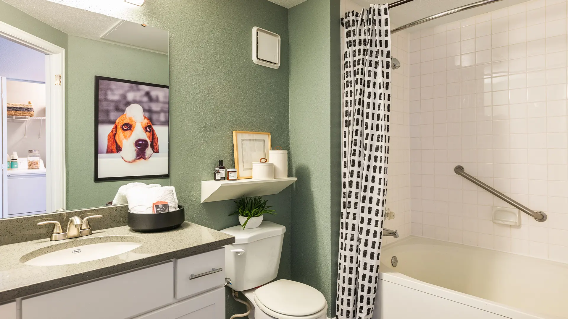 Modern Bathroom with Dog Artwork A modern bathroom featuring a bathtub, a sink with grey countertop, a toilet, and a shower. A framed picture of a dog is on the wall, and there is a plant on a shelf. The walls are painted in muted green tones, with a patterned shower curtain.