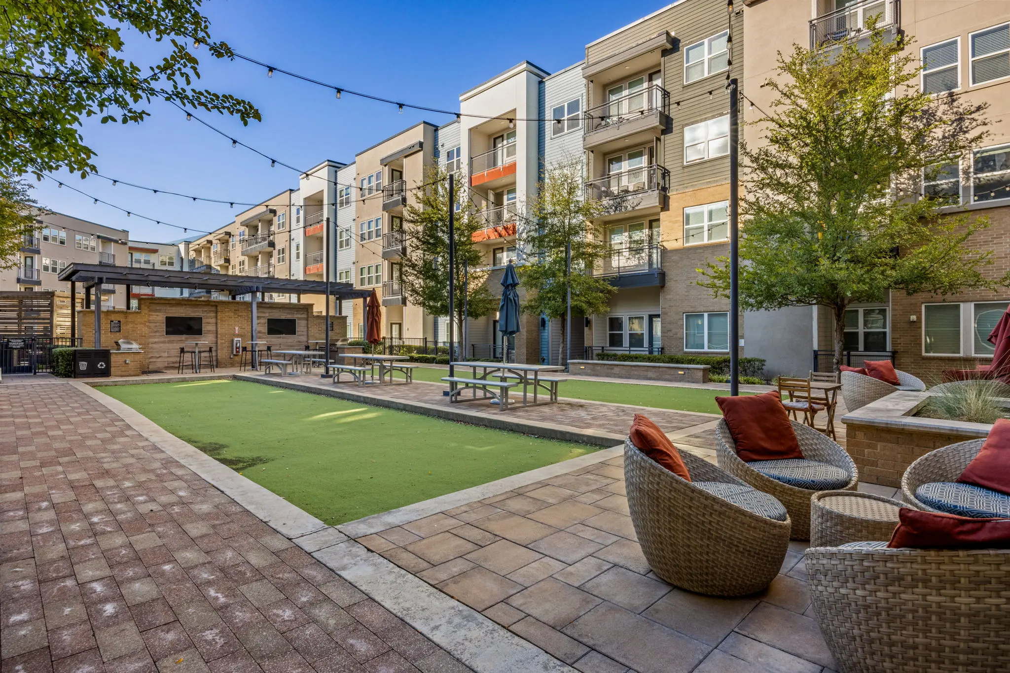 A well-designed outdoor common area in an apartment complex featuring a green lawn space, seating areas with cozy chairs and cushions, and modern buildings in the background.