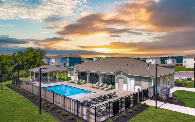 Aerial view of a residential community pool area with loungers and a clubhouse, surrounded by green grass and under a colorful sunset sky.