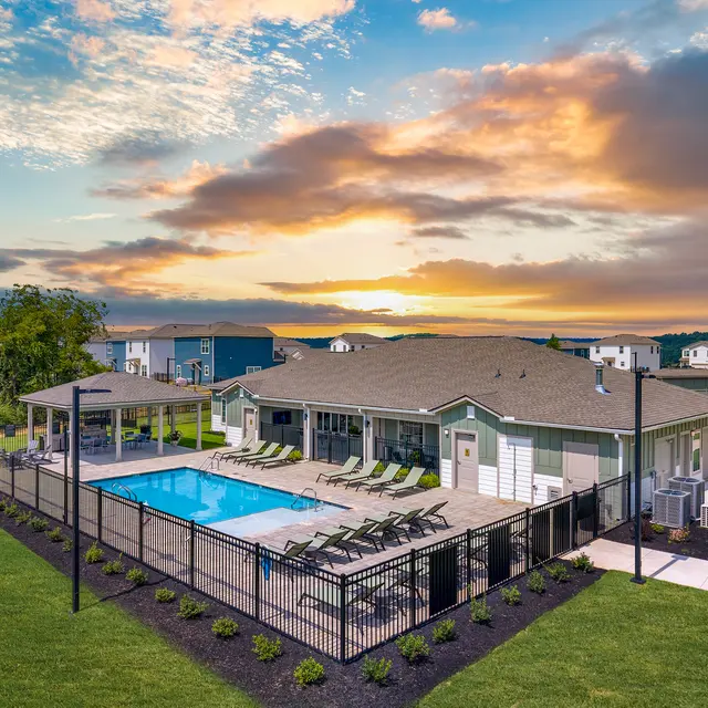 Aerial view of a residential community pool area with loungers and a clubhouse, surrounded by green grass and under a colorful sunset sky.